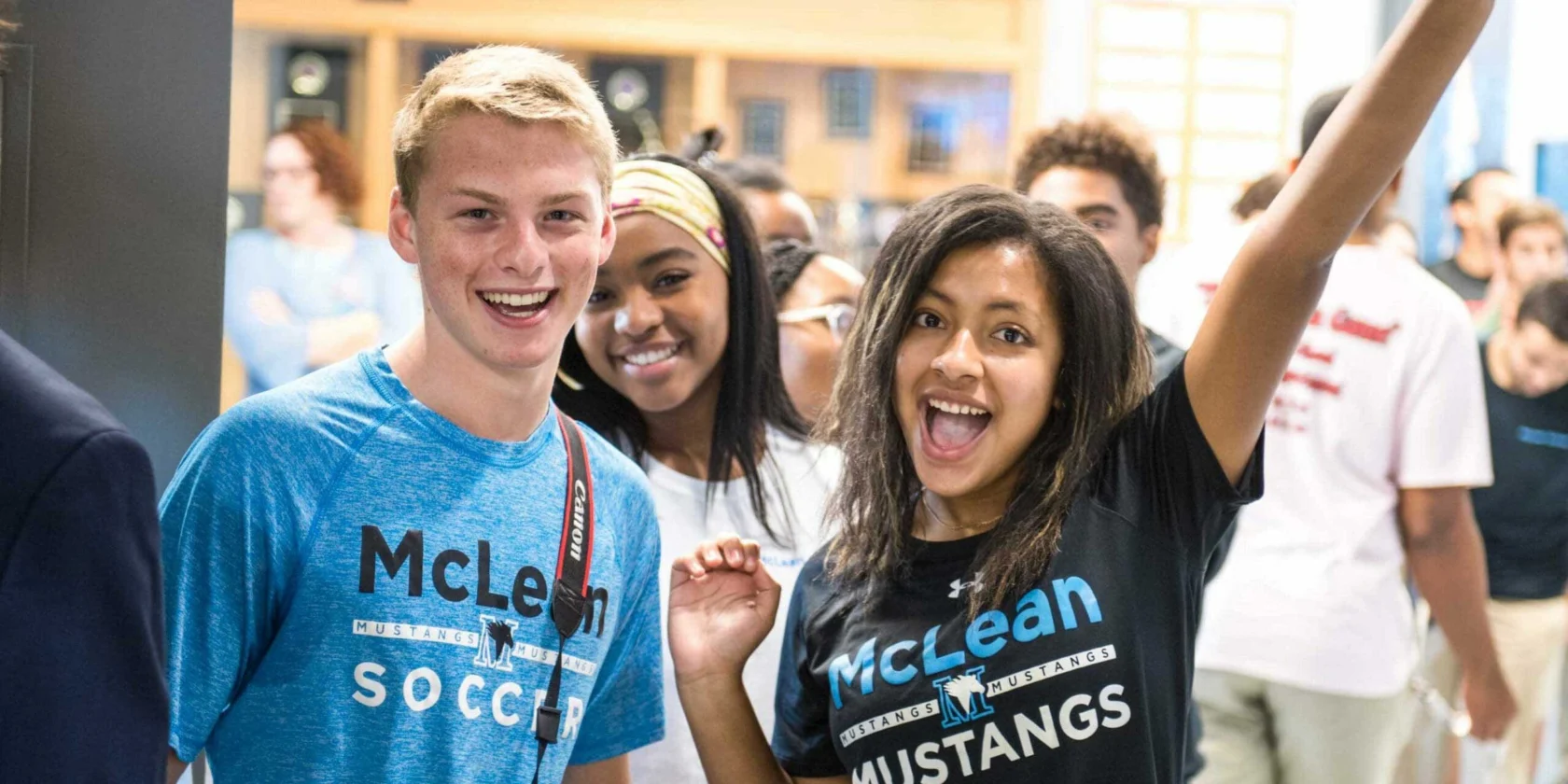 Three students wearing McLean school shirts smile and pose for the camera in a crowded indoor setting; one raises her arm enthusiastically.