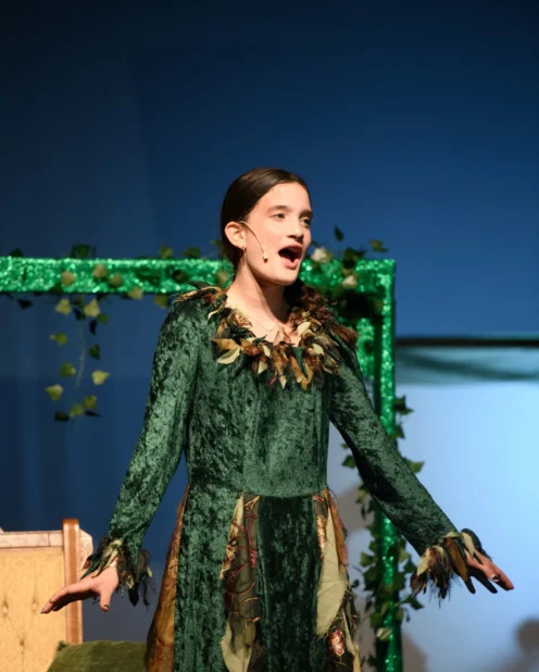 A young person in a green costume performs on stage, wearing a headset microphone, with a decorated green frame and chair in the background.