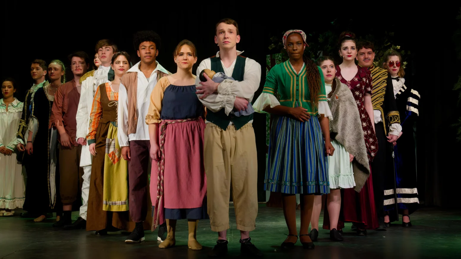 A group of actors in period costumes stand in a line on stage, facing forward, during a theatrical performance.