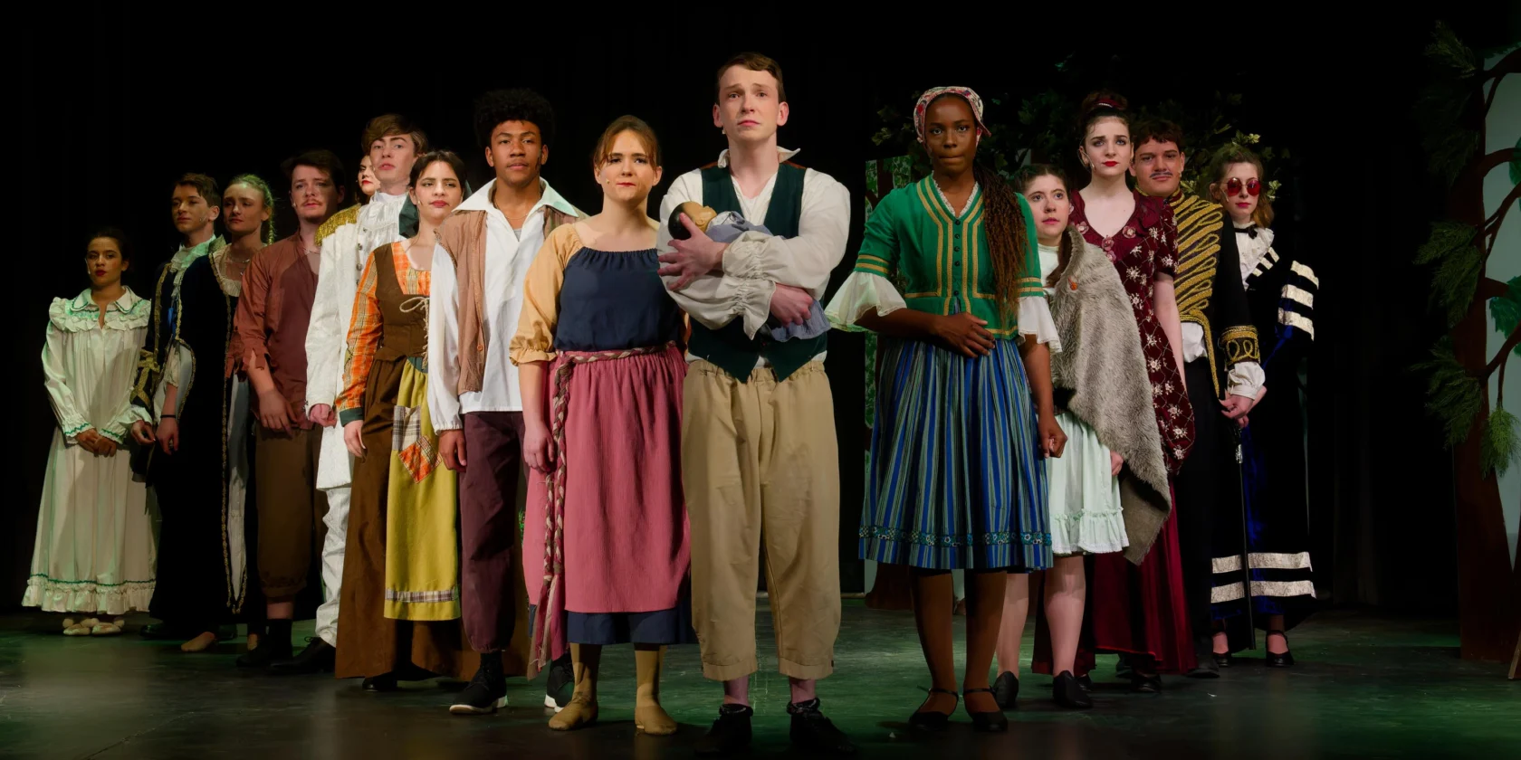 A group of actors in period costumes stand in a line on stage, facing forward, during a theatrical performance.