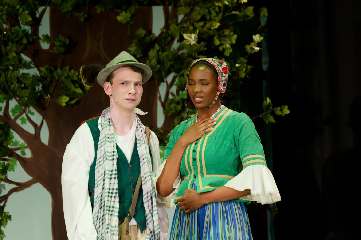 Two actors in period costumes perform on stage in front of a leafy backdrop; one wears a green vest and hat, the other a green top and striped skirt, gesturing with her hand on her chest.