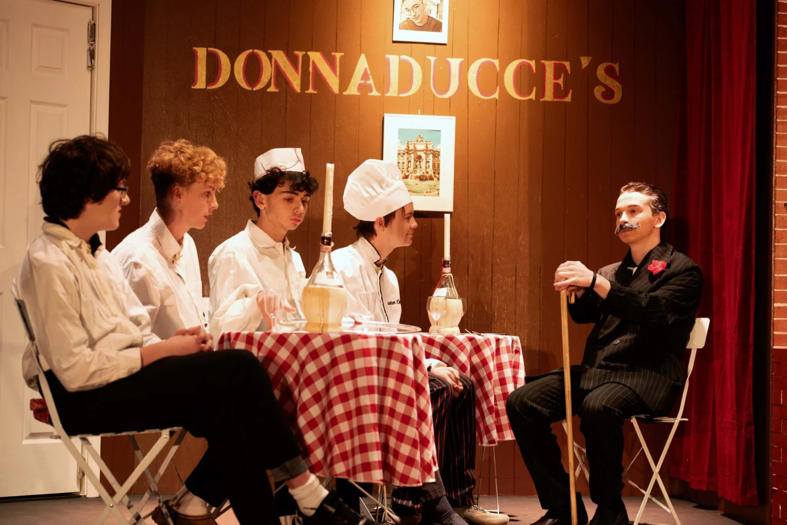 Four people dressed as chefs sit at a table with checkered tablecloths, facing a person in a suit and fake mustache under a sign reading "Donnaducce's.