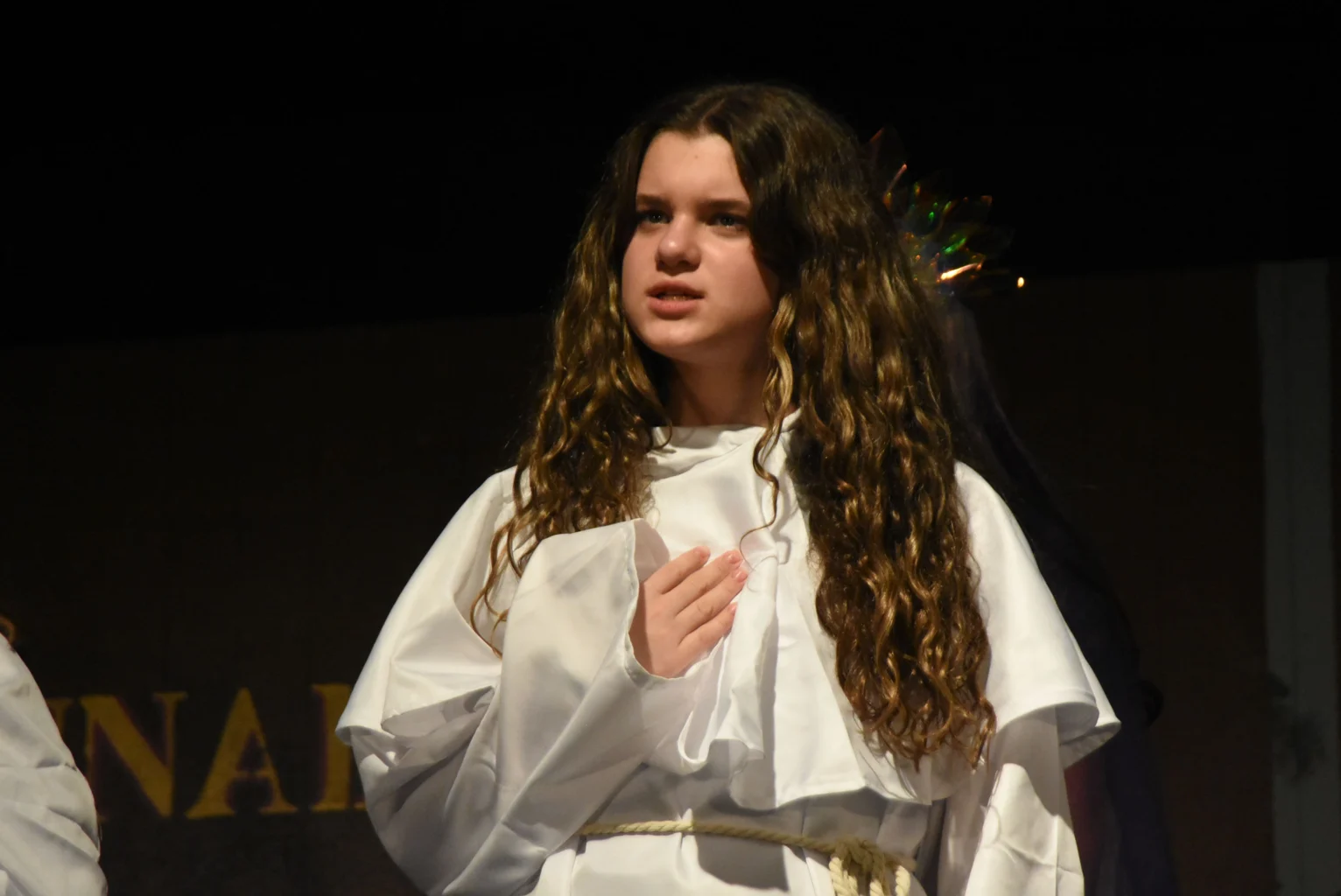 A young person with long curly hair, wearing a white robe, stands with one hand on their chest, appearing to perform on stage.