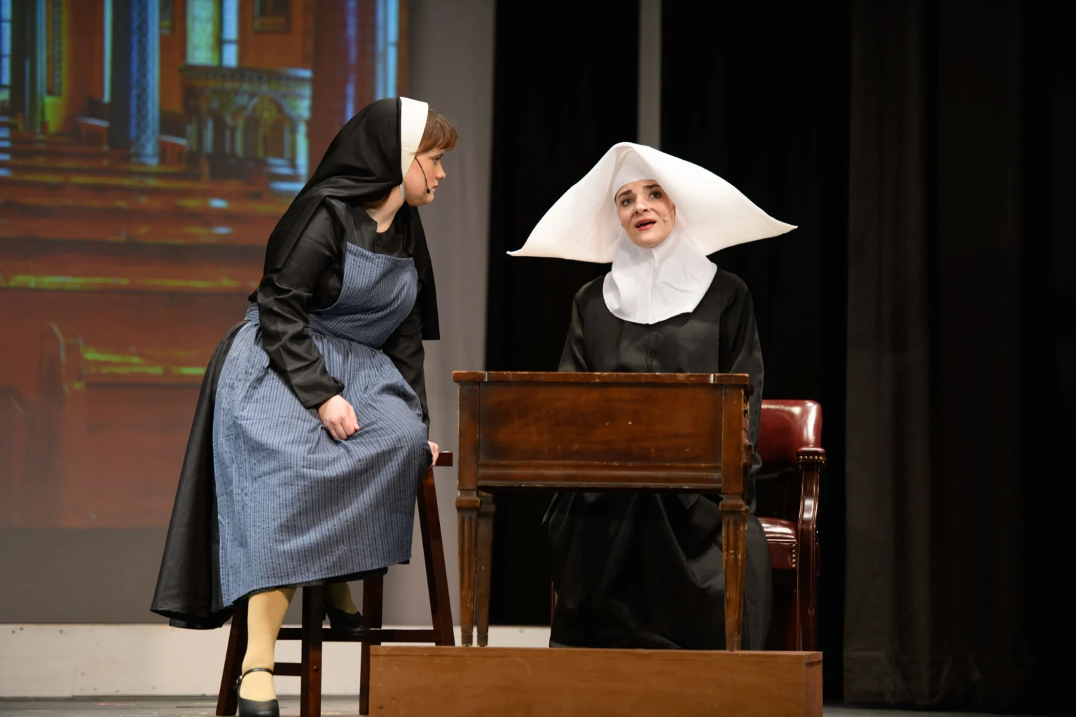 Two women dressed as nuns perform on stage; one stands beside a desk while the other sits behind it, both in period costume with a church background.