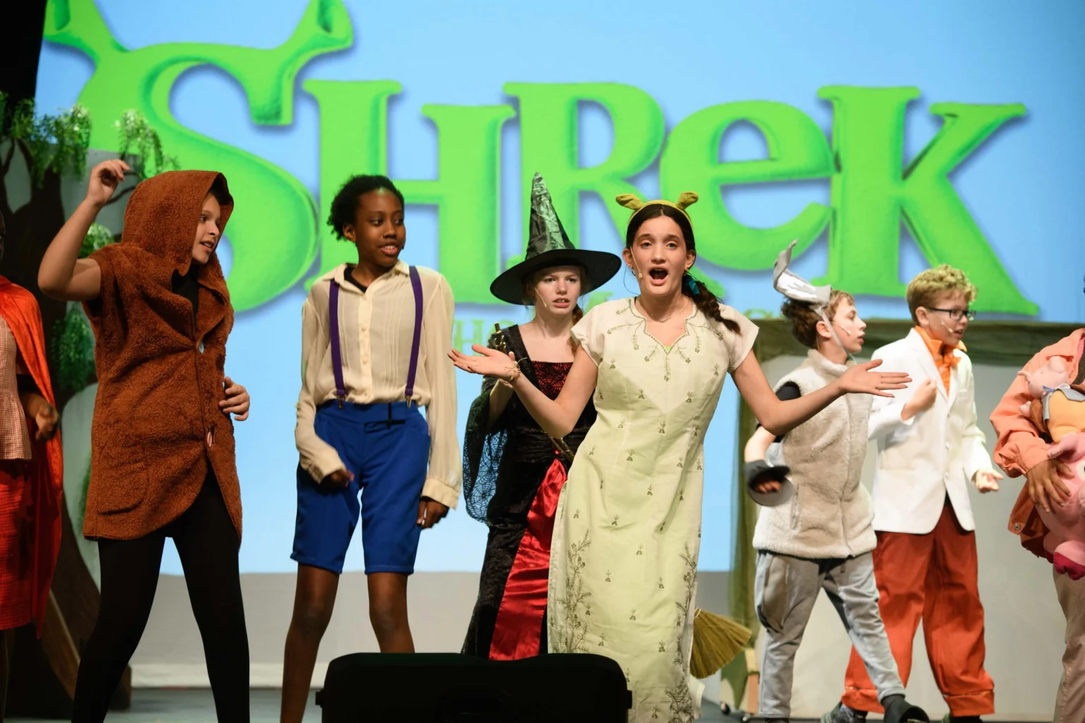 Children in costume perform on stage in front of a large "Shrek" sign, appearing to act out a scene from the musical.
