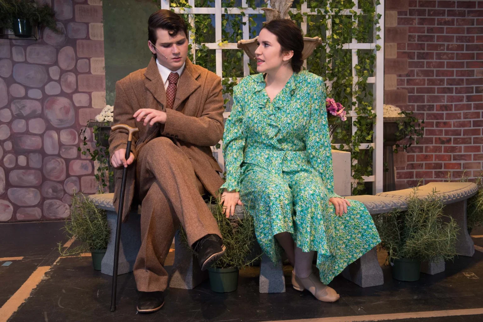 A man in a brown suit with a cane and a woman in a green floral dress sit on a stone bench, engaged in conversation, on a stage with a garden backdrop.