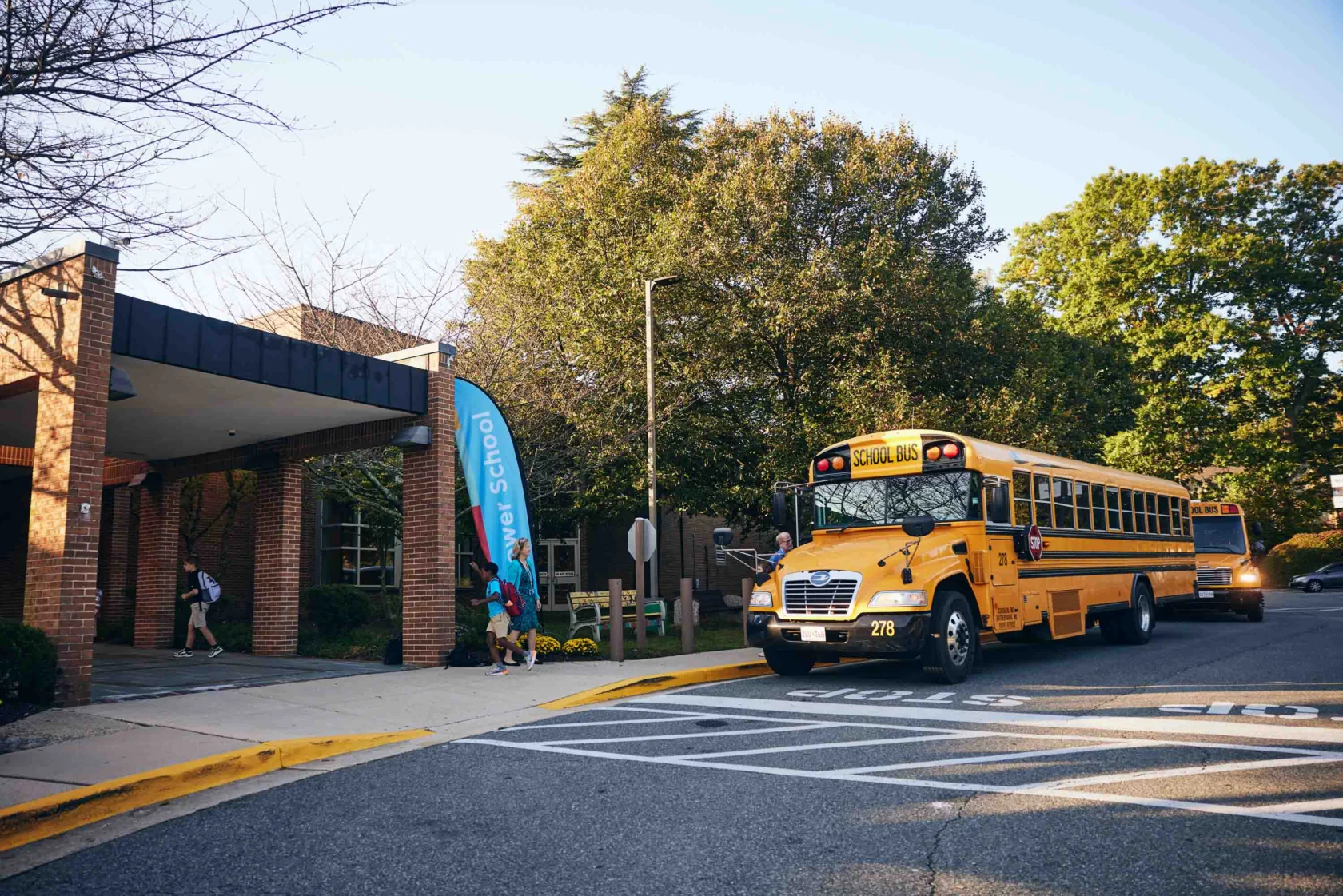 A yellow school bus stops in front of a brick school building as students walk towards the entrance on a sunny day.