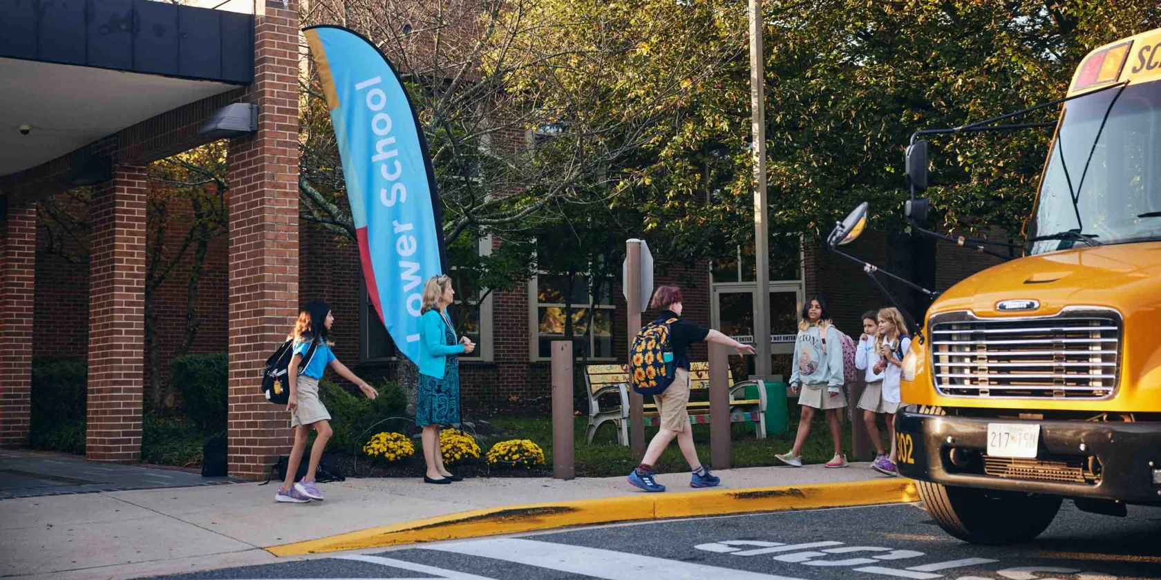 Elementary school students exit a yellow school bus and walk toward a school entrance, as adults greet them near a "Lower School" sign.
