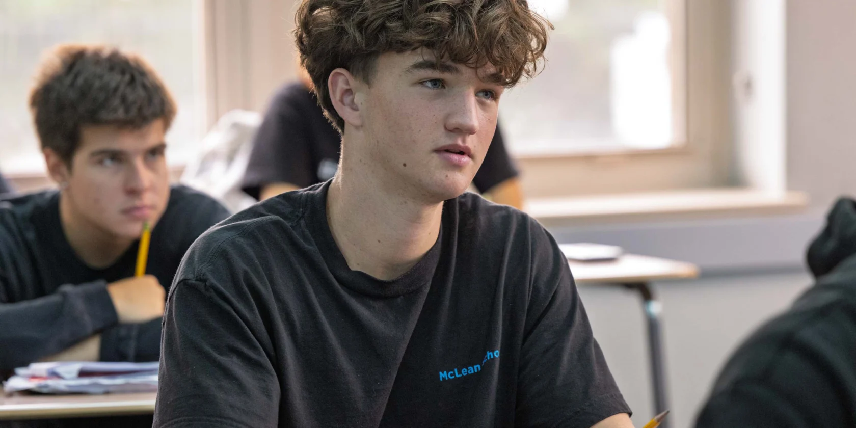 A teenage student with curly hair sits at a desk in a classroom, holding a pencil and looking attentively ahead. Another student is seated behind him.