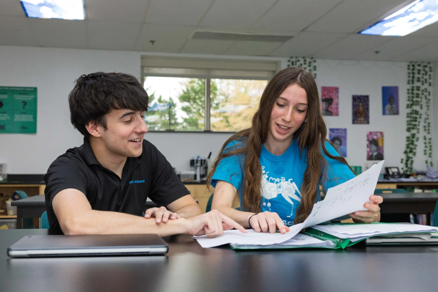 Two students sit at a table in a classroom, looking at and discussing papers together, with a laptop and folders in front of them.