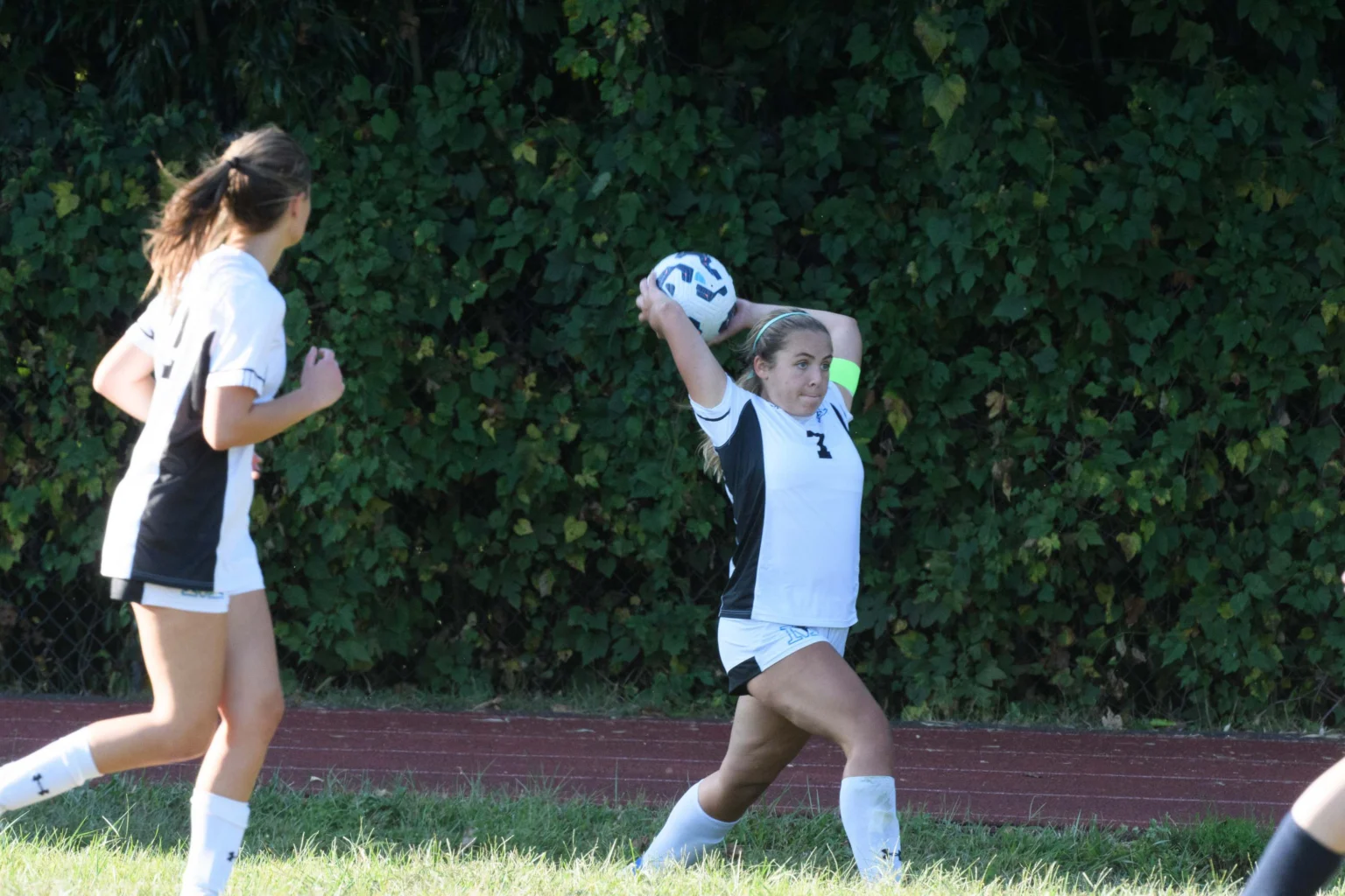 A soccer player prepares to throw the ball from the sideline while a teammate stands nearby on a grassy field with a leafy backdrop.