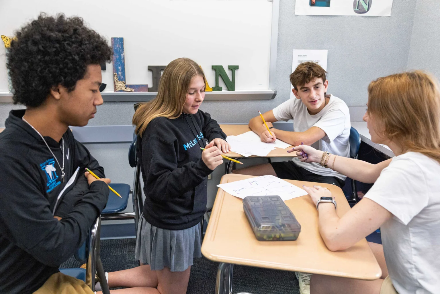 Four students sit around a desk working together on a group project, discussing papers and holding pencils in a classroom setting.