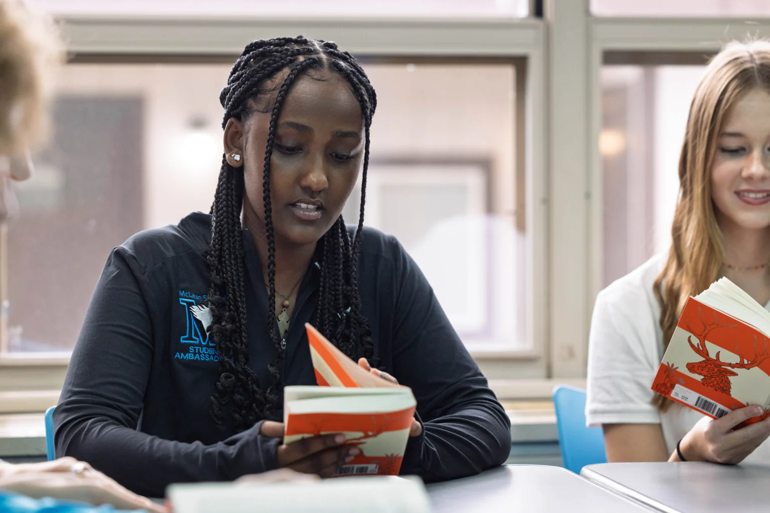 Two students sit at a table reading orange-covered books in a classroom with large windows in the background.