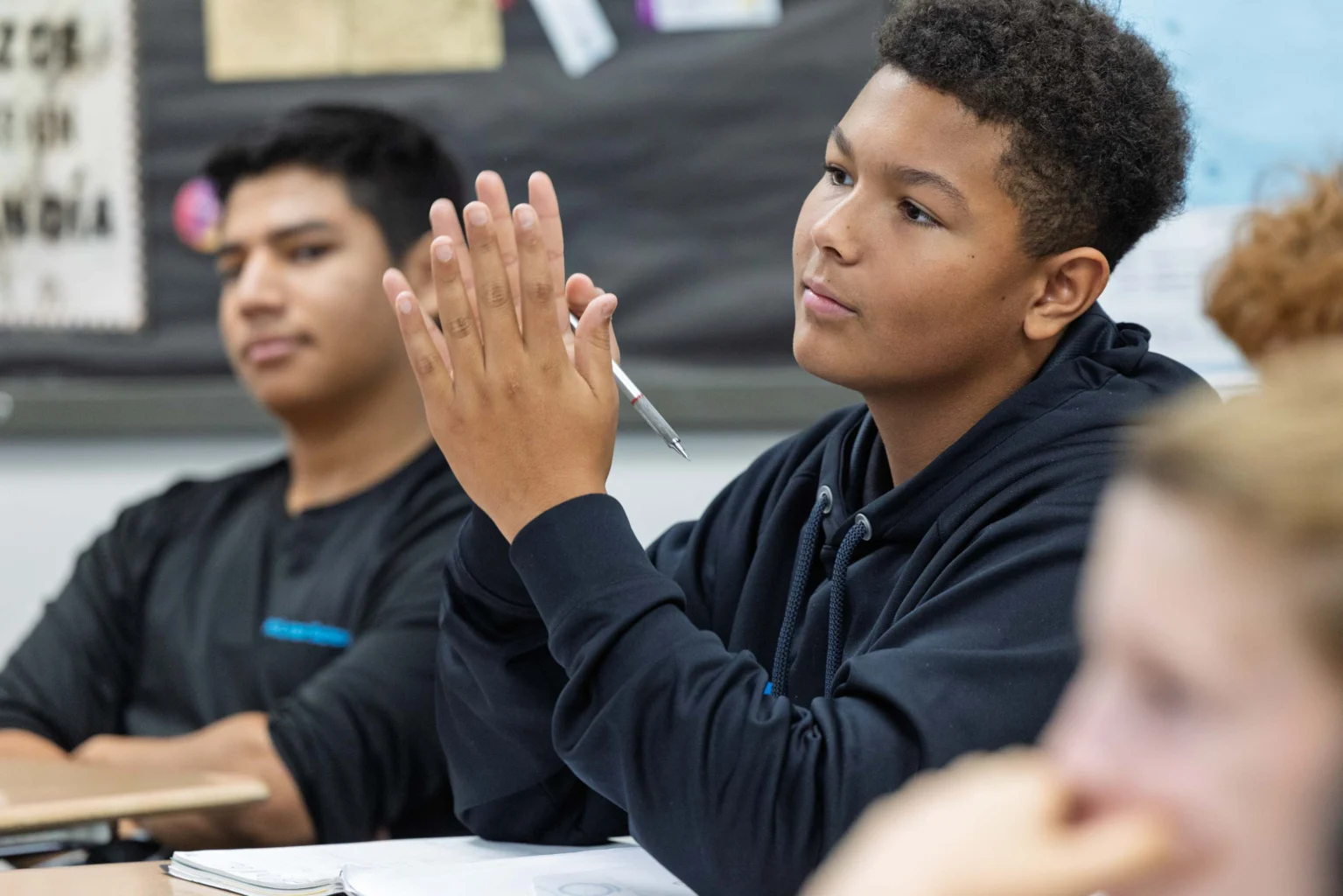 A student in a black hoodie sits at a desk holding a pen and appears attentive in a classroom, with other students visible in the background.