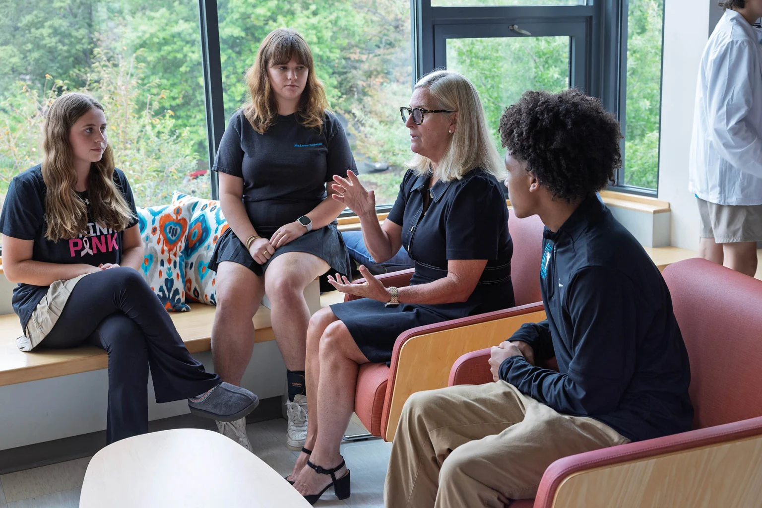 Four people sit and talk in a bright room with large windows; one woman gestures while speaking, and the group listens attentively.
