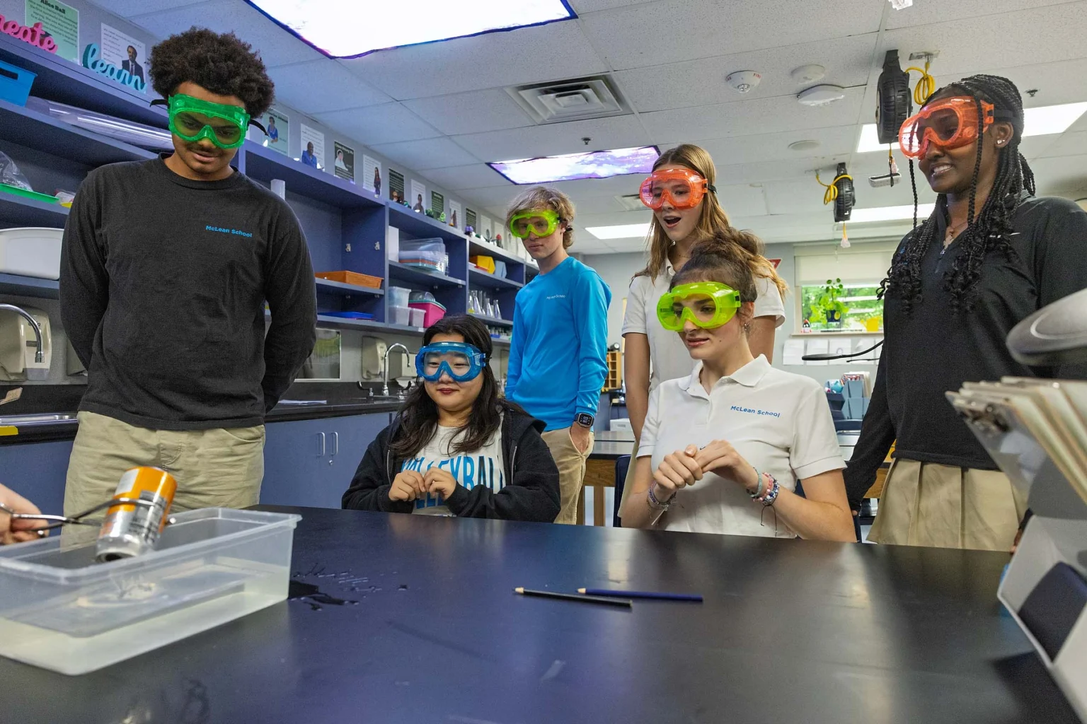 Six students wearing safety goggles observe a science experiment with water at a black lab table in a classroom.