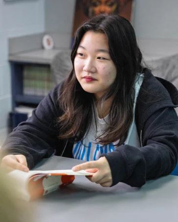 A person with long dark hair sits at a table holding an open book, with shelves of books and a chair visible in the background.