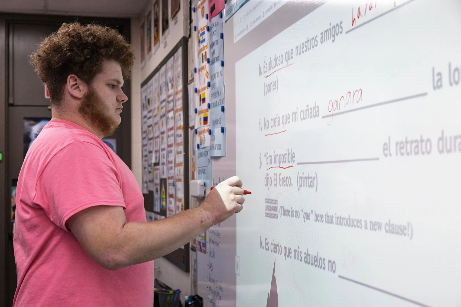 A student in a pink shirt writes answers on an interactive whiteboard displaying Spanish grammar exercises.