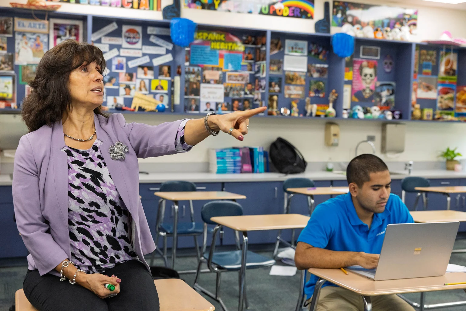 A teacher sits on a desk and gestures while speaking, as a student types on a laptop in a colorful classroom.