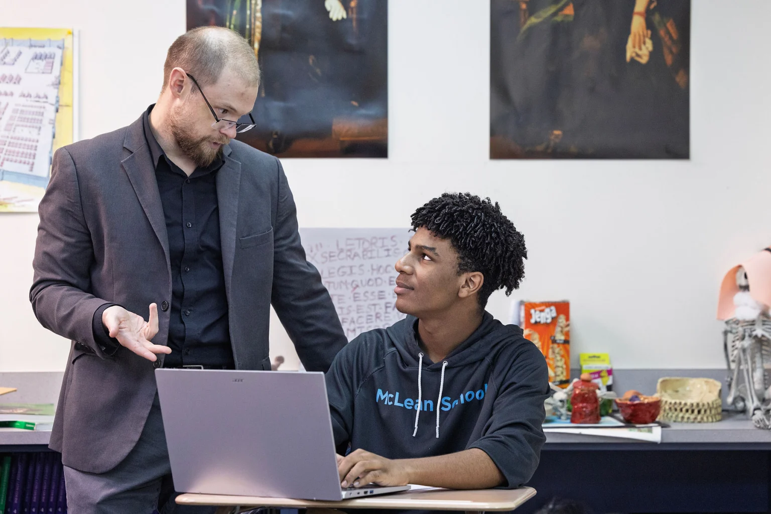 A teacher in a suit talks to a student sitting at a desk with a laptop in a classroom decorated with posters and educational materials.