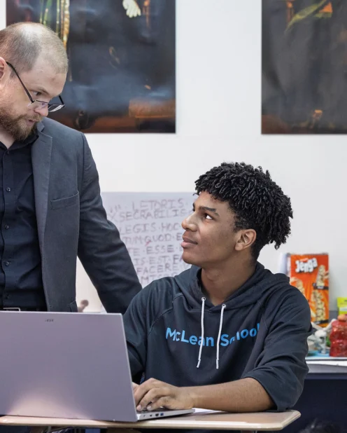 A teacher in a suit talks to a student sitting at a desk with a laptop in a classroom decorated with posters and educational materials.