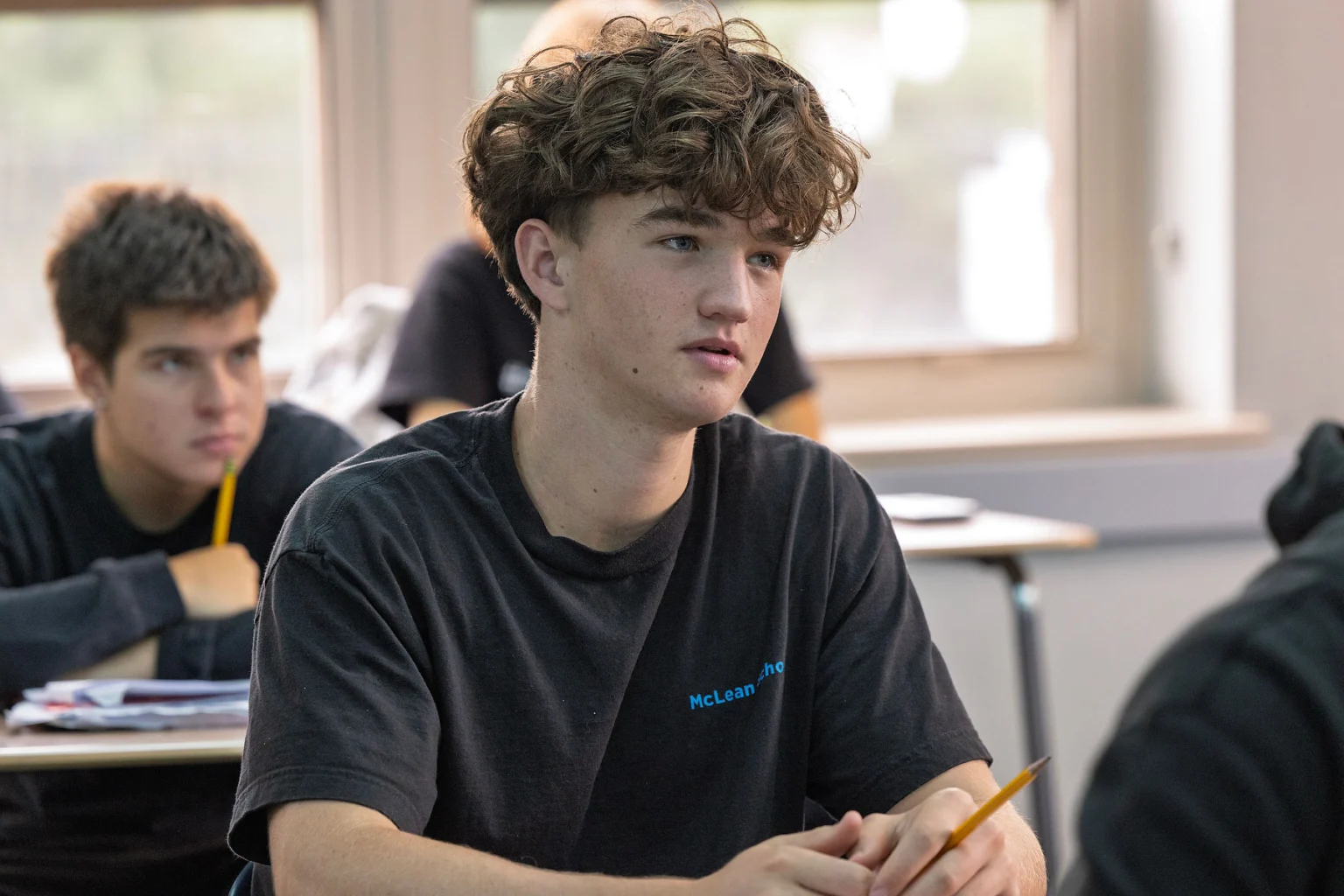 A teenage student with curly hair sits at a desk in a classroom, holding a pencil and looking attentive. Another student is visible in the background.