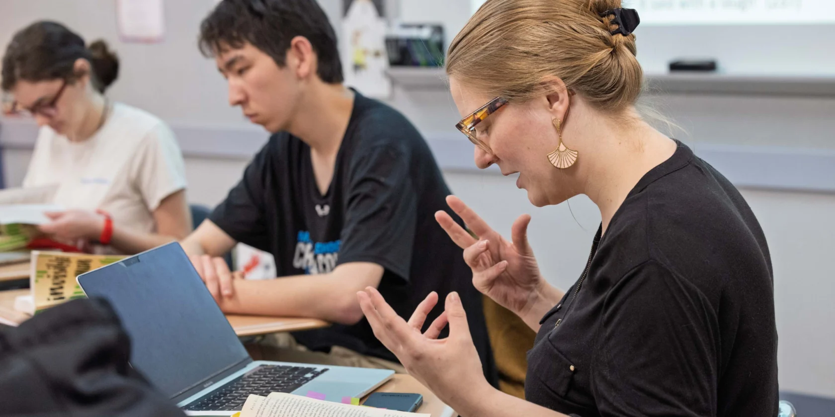 A teacher gestures while speaking to students in a classroom; books and a laptop are open on the desk, and projected text is visible on the board in the background.