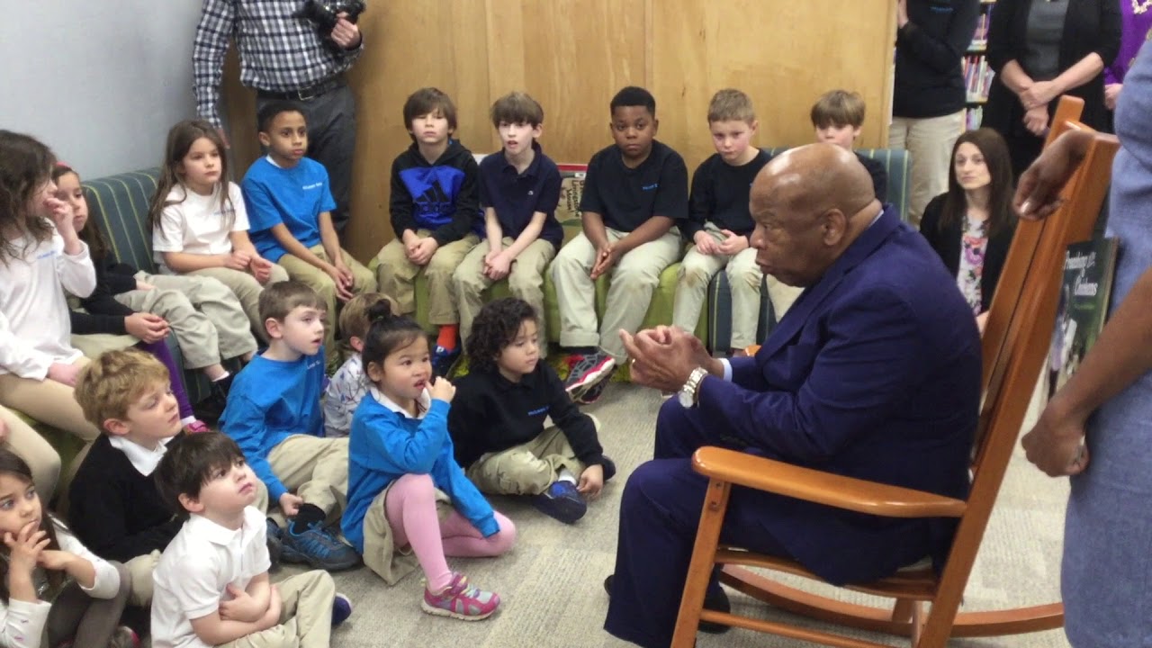 An older man in a suit sits in a rocking chair, speaking to a group of children seated on the floor around him in a classroom or library setting.