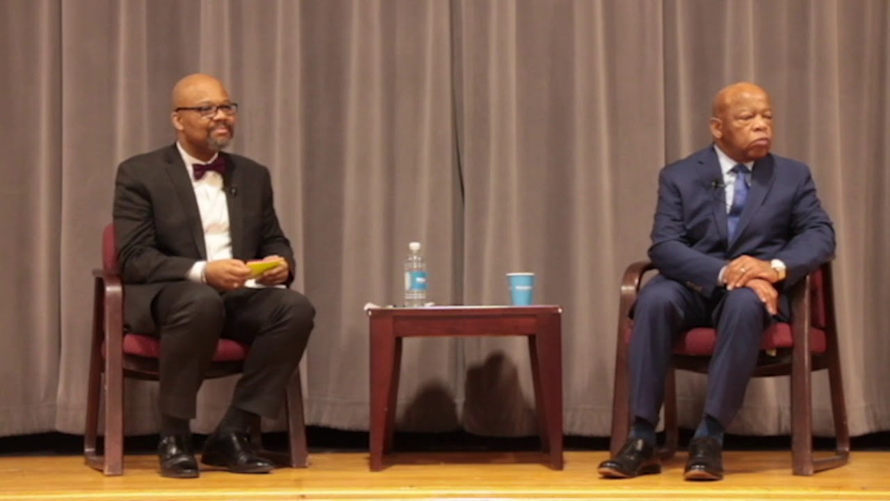Two men in suits sit on stage in chairs, separated by a small table with a water bottle, blue cup, and papers, against a backdrop of closed gray curtains.