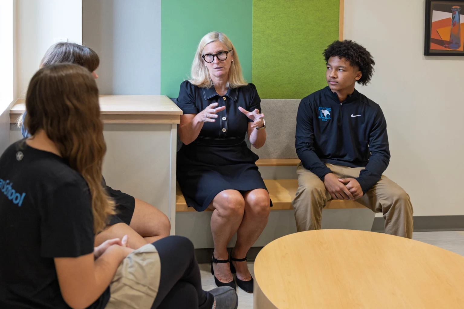 A woman in a black dress is speaking to three students seated around her in a modern classroom setting.