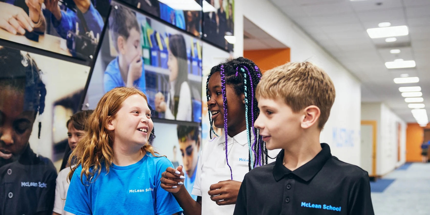 Three children in McLean School uniforms talk and smile in a hallway with student photos on the wall in the background.