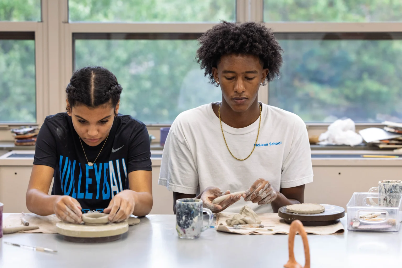 Two students sit at a table working with clay on pottery wheels in a classroom, focused on shaping their pieces.