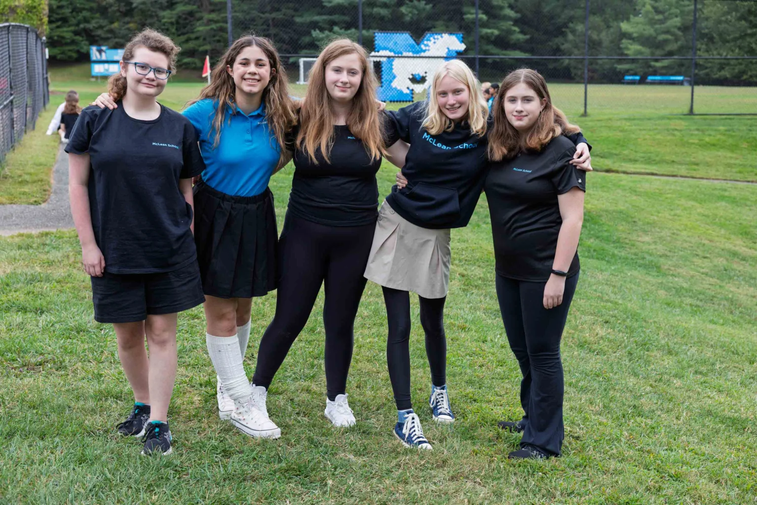 Five teenage girls stand together on a grassy field, posing for a group photo. Four wear black shirts, one wears a blue shirt. A chain-link fence and trees are visible in the background.