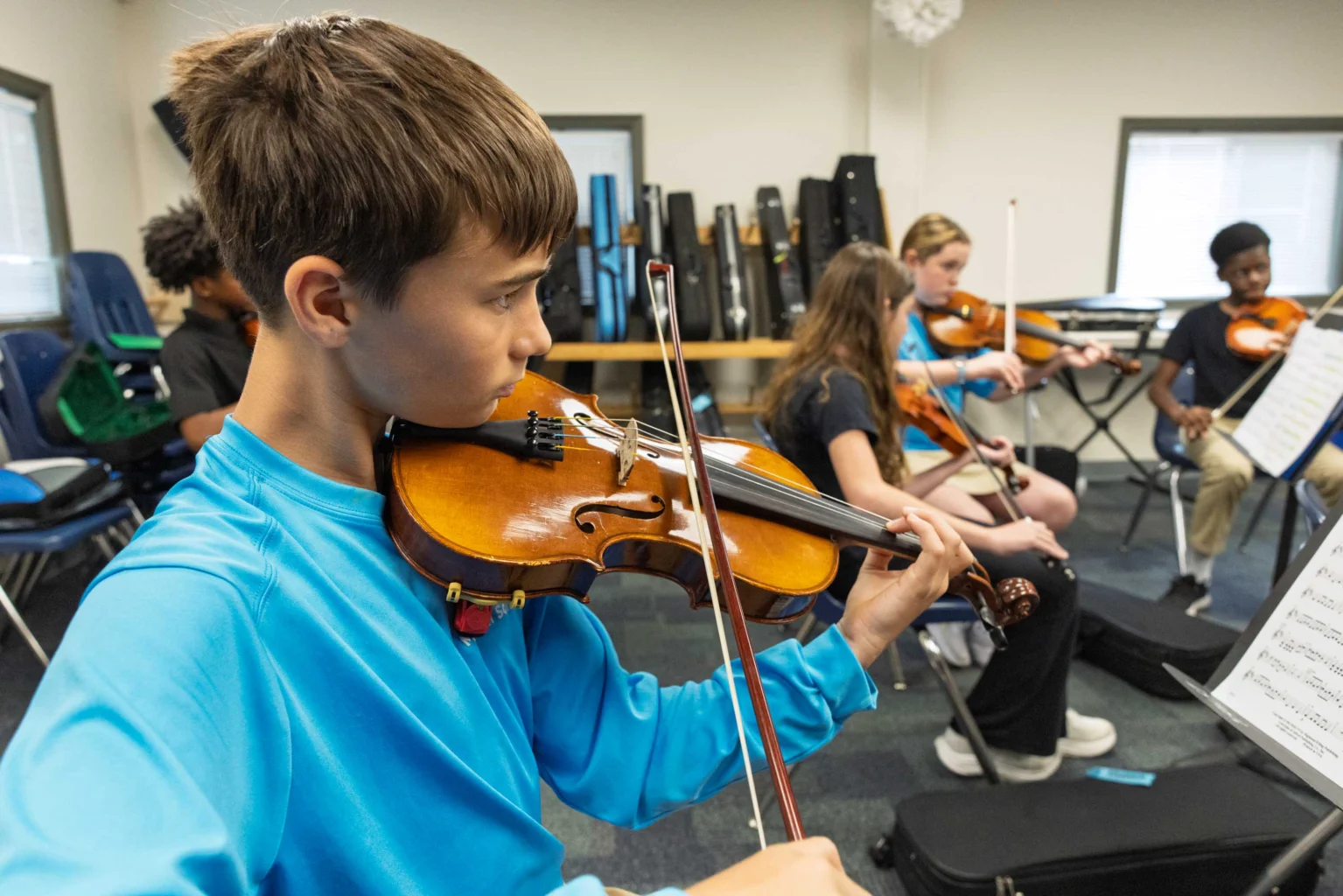 A boy in a blue shirt plays the violin in a classroom with other students, who are also playing string instruments and reading sheet music.