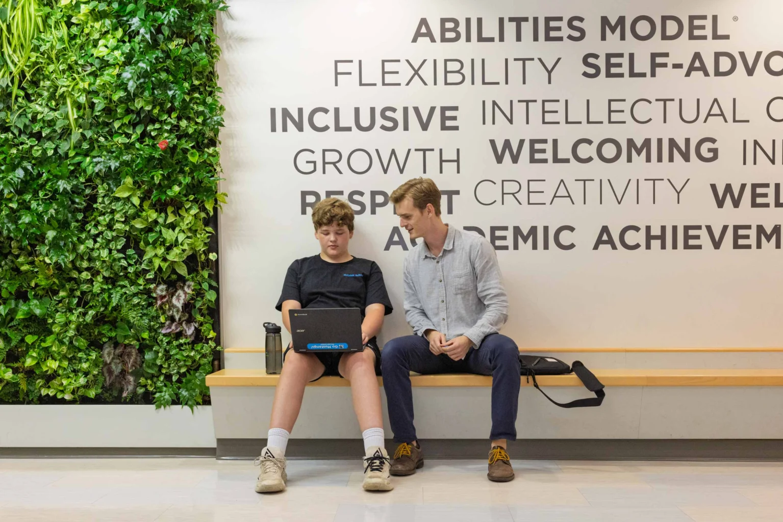 Two people sit on a wooden bench; one uses a laptop while the other looks on. A plant wall and motivational words are visible in the background.