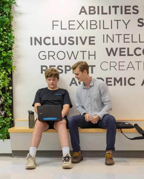 Two people sit on a wooden bench; one uses a laptop while the other looks on. A plant wall and motivational words are visible in the background.
