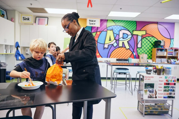 An adult assists a child with an art project in a colorful classroom labeled "ART," with supplies and artwork visible in the background.