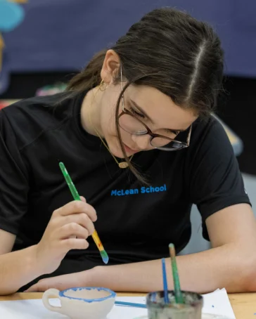 A student wearing glasses and a McLean School shirt paints a ceramic bowl at a classroom table. Art supplies are visible around her.