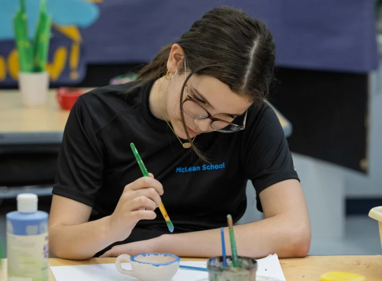 A student wearing glasses and a McLean School shirt paints a ceramic bowl at a classroom table. Art supplies are visible around her.