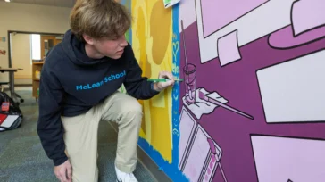A student wearing a "McLean School" hoodie kneels while painting a mural with purple, yellow, and blue sections on a classroom wall.