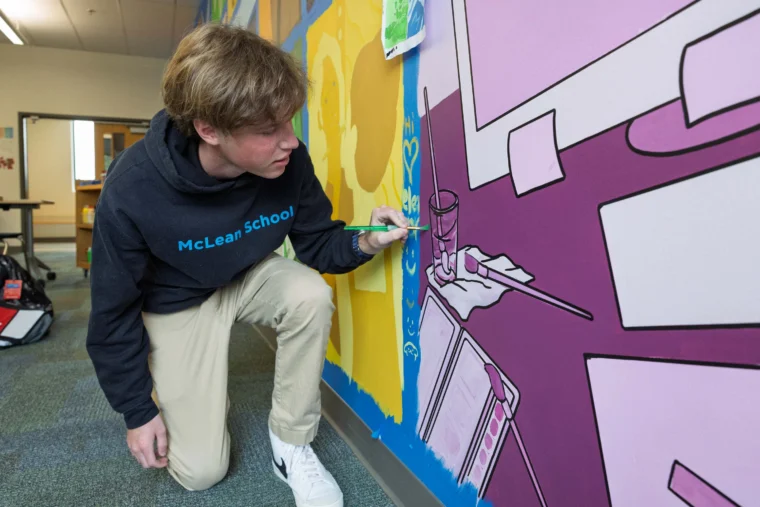 A student wearing a "McLean School" hoodie kneels while painting a mural with purple, yellow, and blue sections on a classroom wall.