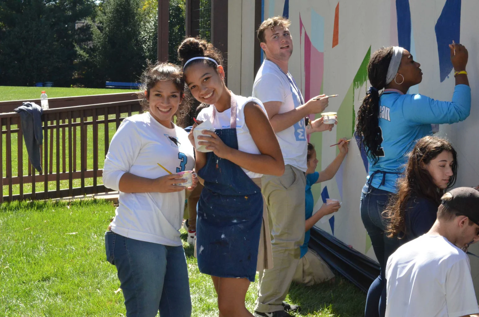 A group of young adults paint a colorful mural on a large outdoor wall on a sunny day; some are smiling at the camera while others focus on painting.