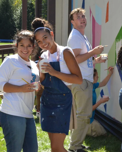 A group of young adults paint a colorful mural on a large outdoor wall on a sunny day; some are smiling at the camera while others focus on painting.