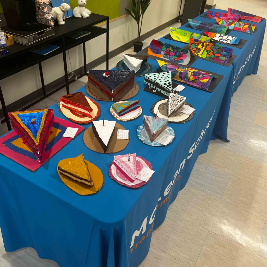 A display of colorful, geometric cake slice models made from various materials arranged on a blue tablecloth in a classroom setting.