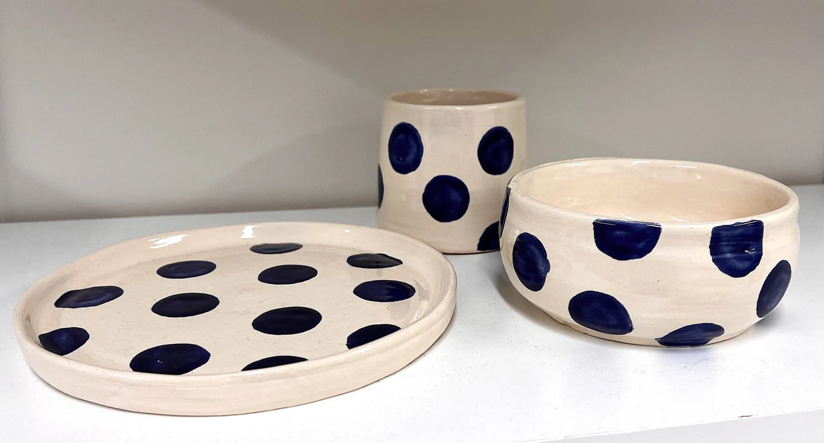 Three handmade ceramic pieces—a plate, a tumbler, and a bowl—each decorated with large blue polka dots, displayed on a white shelf.