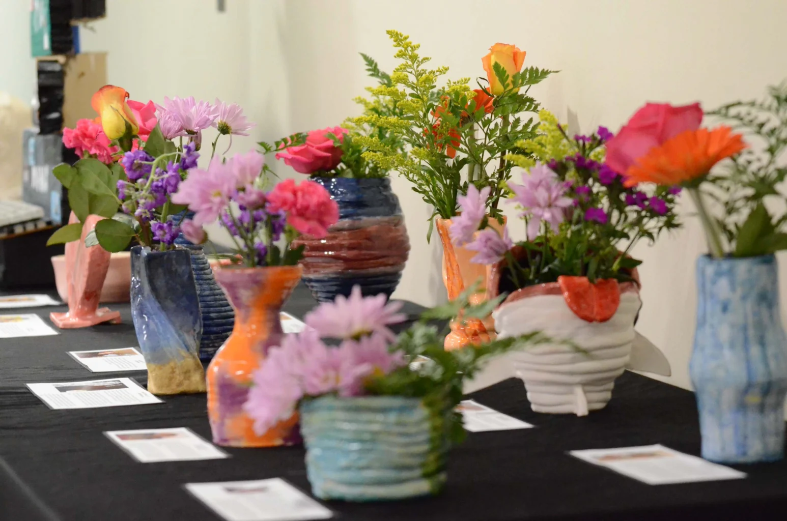 Several colorful ceramic vases filled with various bright flowers are displayed on a black table, each vase accompanied by a printed information card.