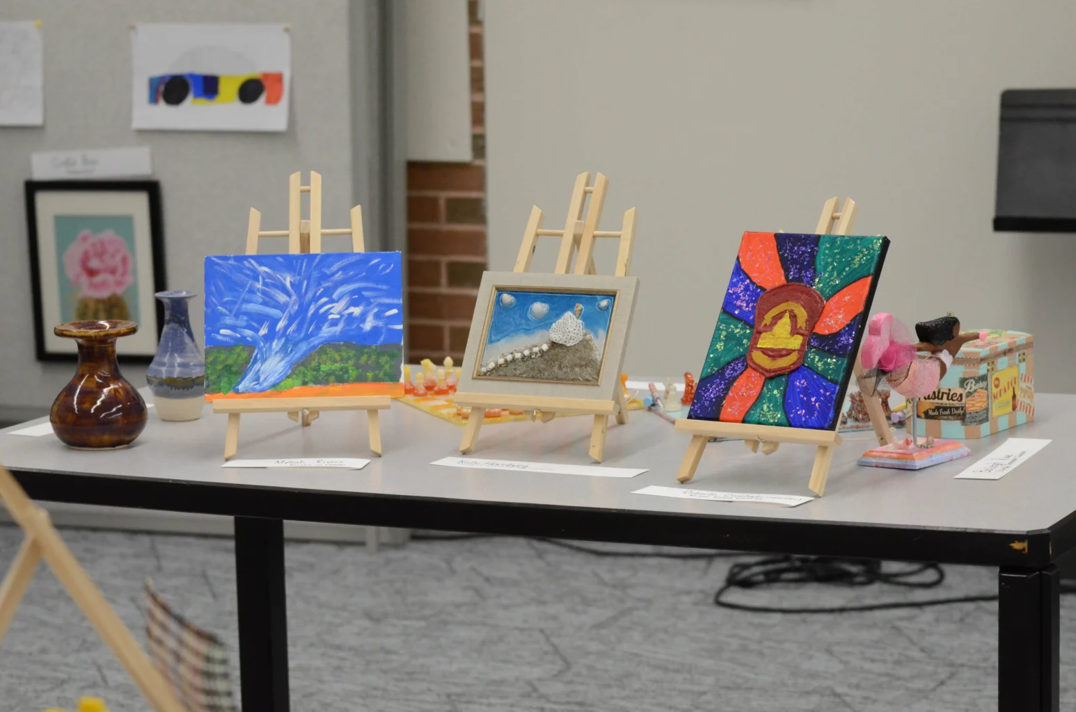 Three small paintings on easels are displayed on a table, with pottery, a framed photo, and other art items in the background.