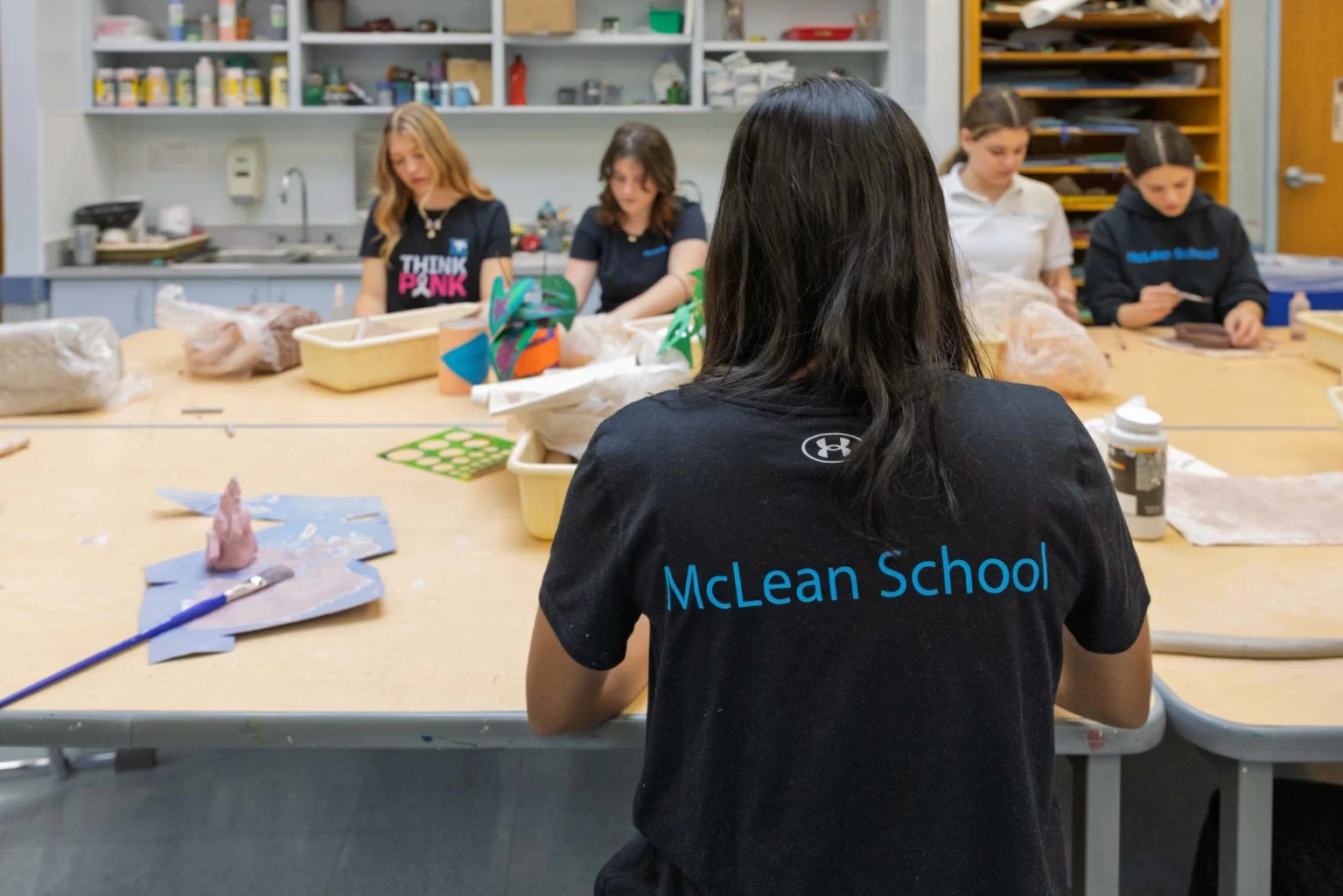 Students sit around a table working on art projects in a classroom; one student in the foreground wears a shirt with “McLean School” on the back. Shelves with supplies are visible behind them.