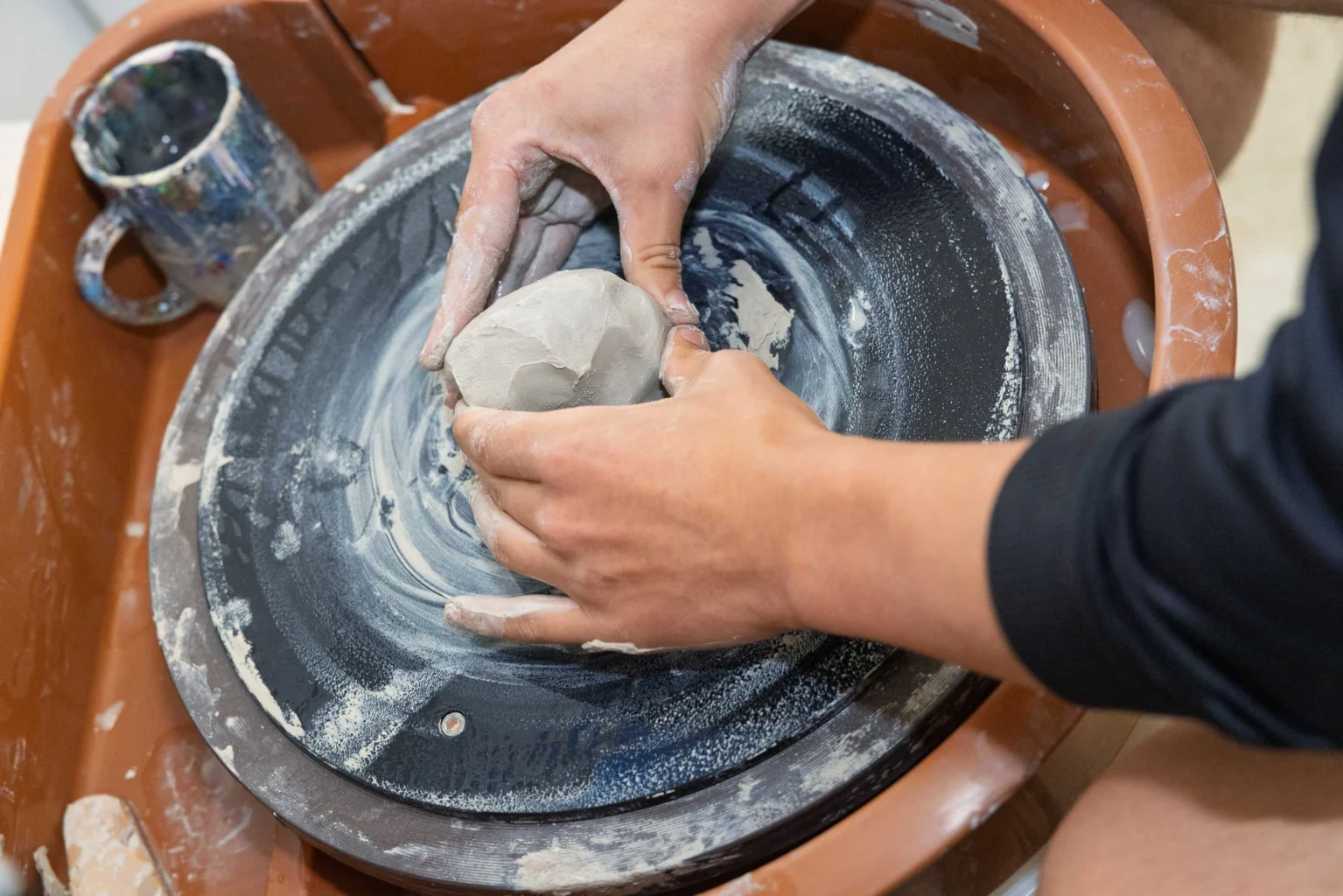 Hands shaping a ball of wet clay on a pottery wheel, with a ceramic cup nearby and clay residue visible on the wheel’s surface.