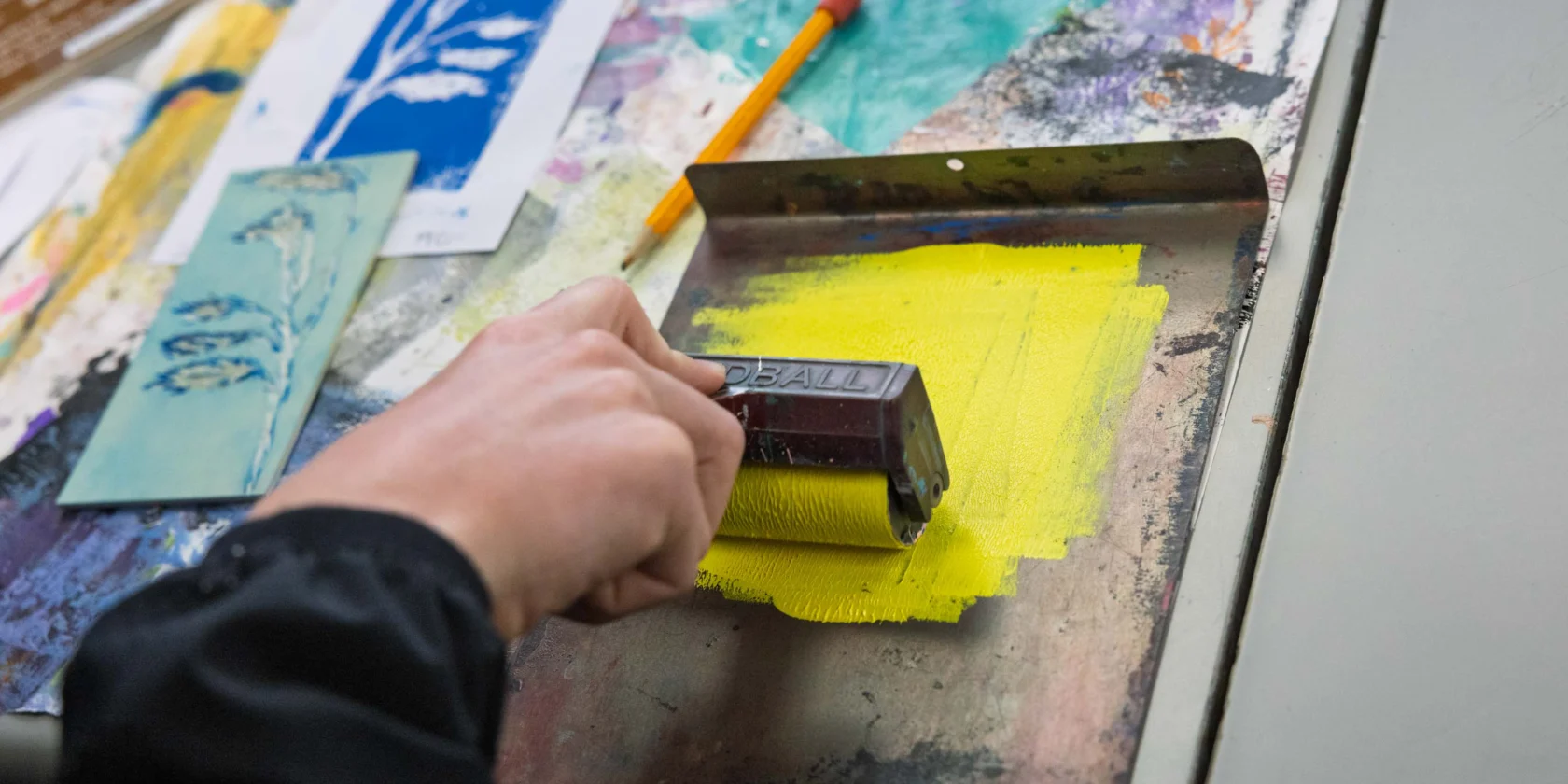 A person uses a brayer to roll yellow ink onto a metal surface, with art supplies, paper, and painted textures visible on the table.
